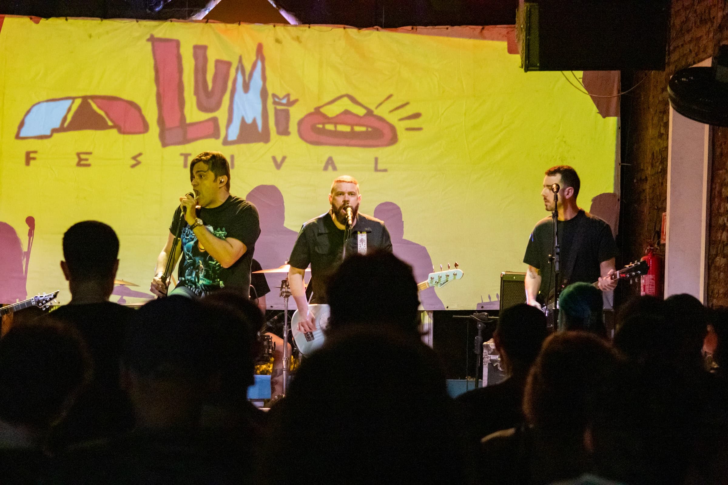 Banda toca diante da marca Alumiô projetada em amarelo, com a plateia próxima ao palco.