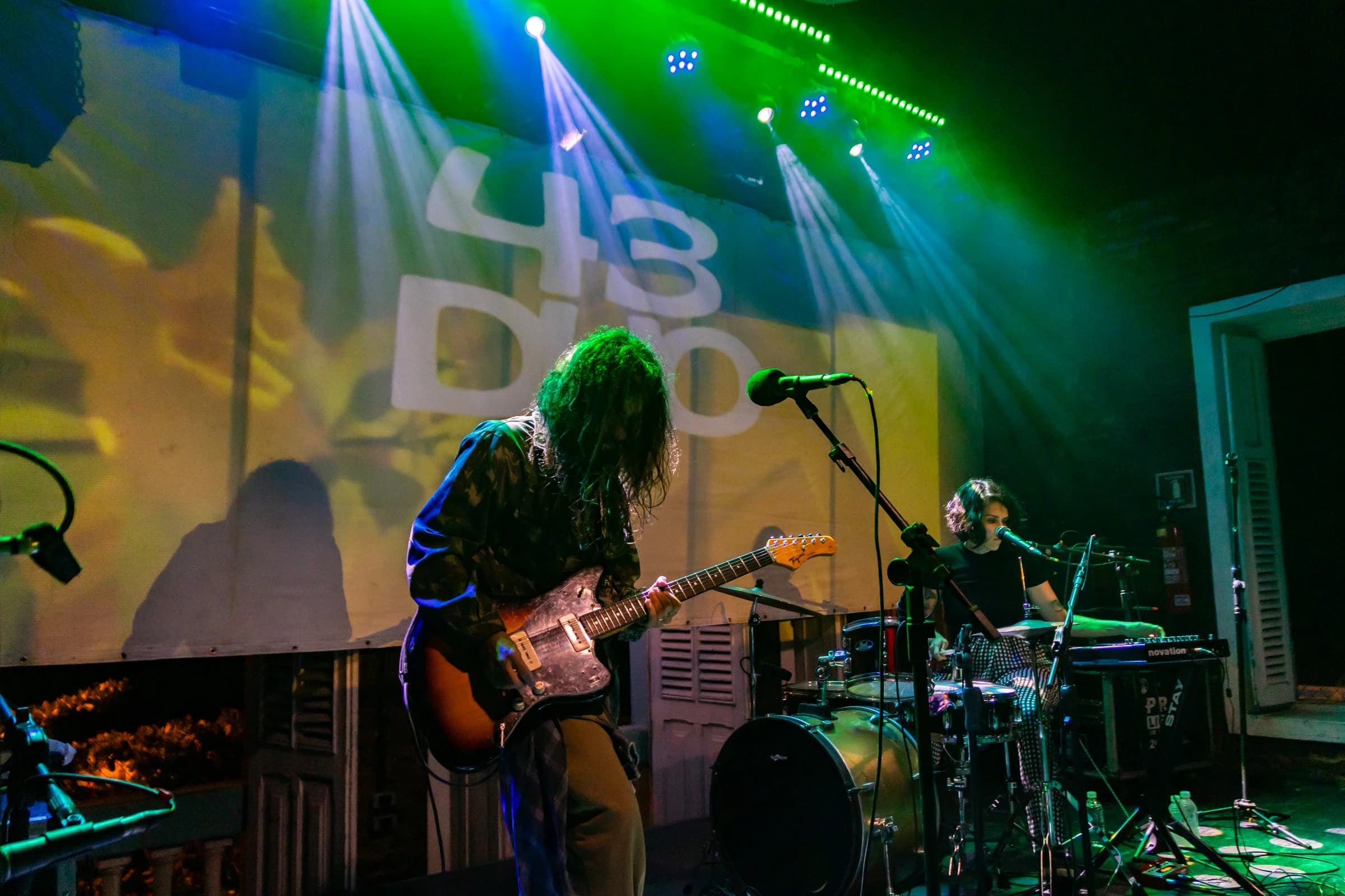 Guitarrista de cabelos longos e baterista se apresentam sob luzes verdes e amarelas.