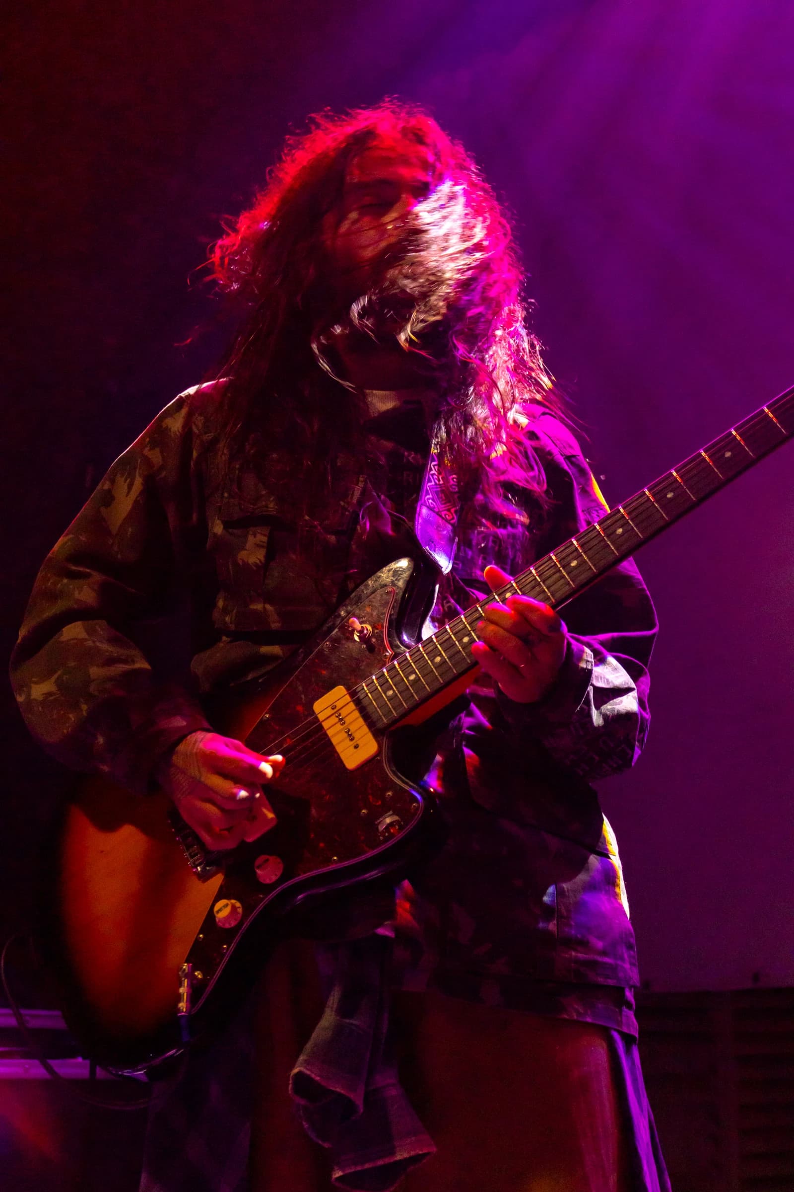 Guitarrista de cabelos longos toca sob iluminação magenta.