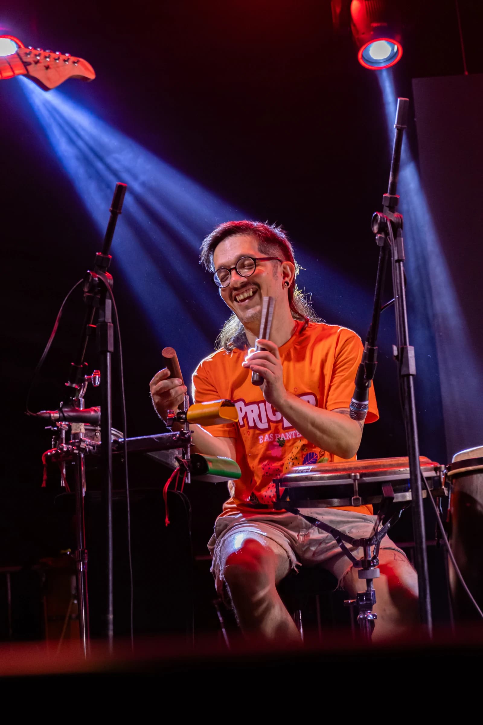 Percussionista de óculos e camiseta laranja sorri durante a apresentação.