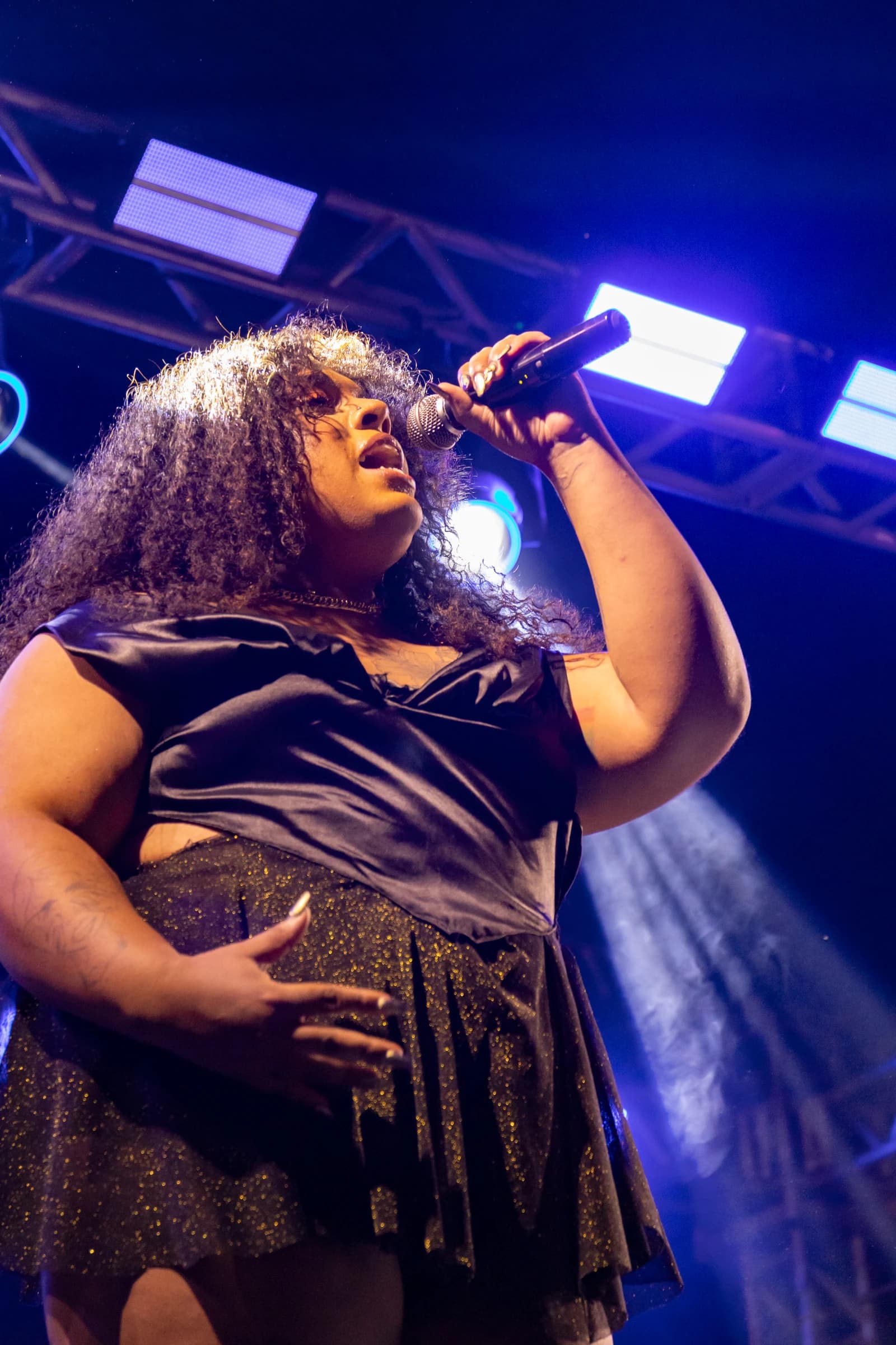 Vocalista de roupa preta canta ao microfone, vista de baixo contra as luzes do palco.